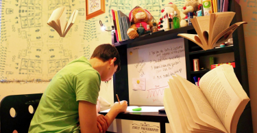 male college student in green shirt writing a paper at his desk
