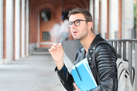 student wearing glasses sitting outside of school smoking while holding his books