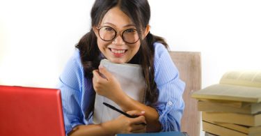 young sweet and happy Asian Chinese student girl in nerd glasses working cheerful on laptop computer on desk with pile of books studying and smiling isolated on white background