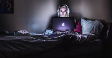 blonde college student on her laptop in the dark in her bed