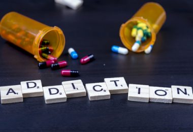 scrabble letters laid out on table spelling out the word addiction with pills spilled out of their containers in the background.