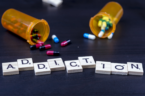 scrabble letters laid out on table spelling out the word addiction with pills spilled out of their containers in the background.