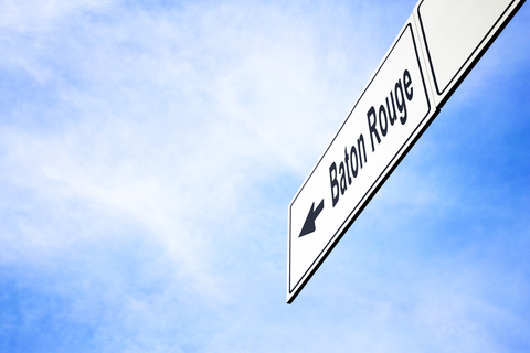 White signboard with an arrow pointing left towards Baton Rouge, Louisiana, USA, against a hazy blue sky in a concept of travel, navigation and direction. Path included for the signboard