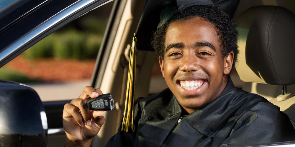 young male college graduate sitting in his first car holding his keys and smiling