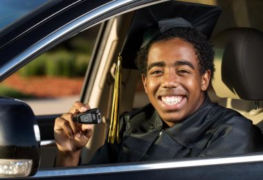 young male college graduate sitting in his first car holding his keys and smiling