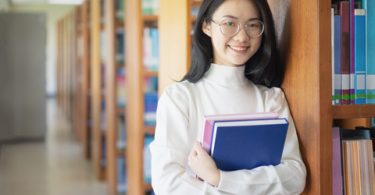 smiling female asian college student holding her books and standing in the library