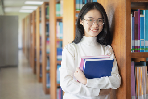 smiling female asian college student holding her books and standing in the library