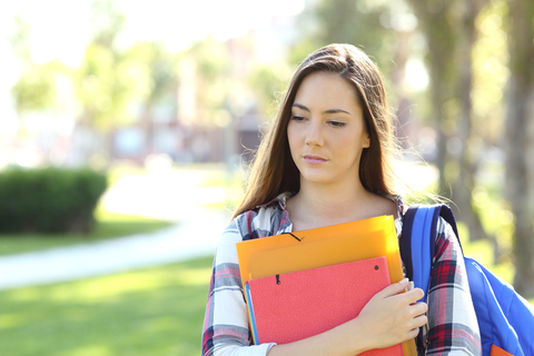 Front view portrait of a sad student walking in the street holding folders outdoors in a park