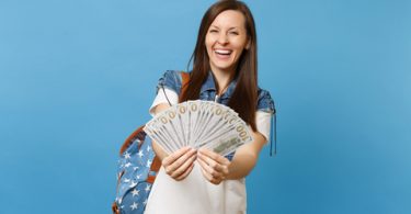 Portrait of female college student laughing in denim clothes with backpack holding bundle lots of dollars