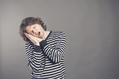Male college student in striped black and white sweater wants to sleep. Put his head in his hands and closed eyes while yawning. Standing on the grey background.