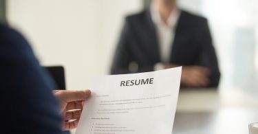 employer holding up resume at desk, interviewee sitting in the background