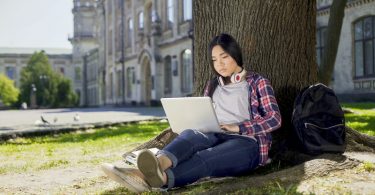 College student sitting under tree in campus, using laptop doing online course