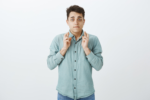 male student standing in front of white background crossing fingers looking nervous
