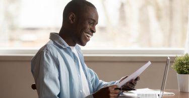 African guy sitting at table reading college acceptance letter with good news