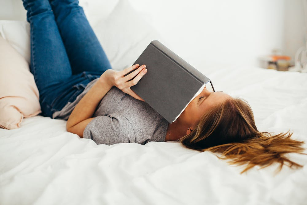 female college student laying on bed with book covering her face