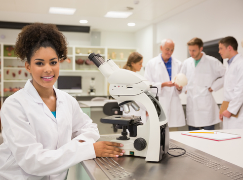 female college student smiling in lab coat sitting next to microscope