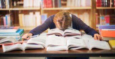 College student laying head on pile of text books on desk